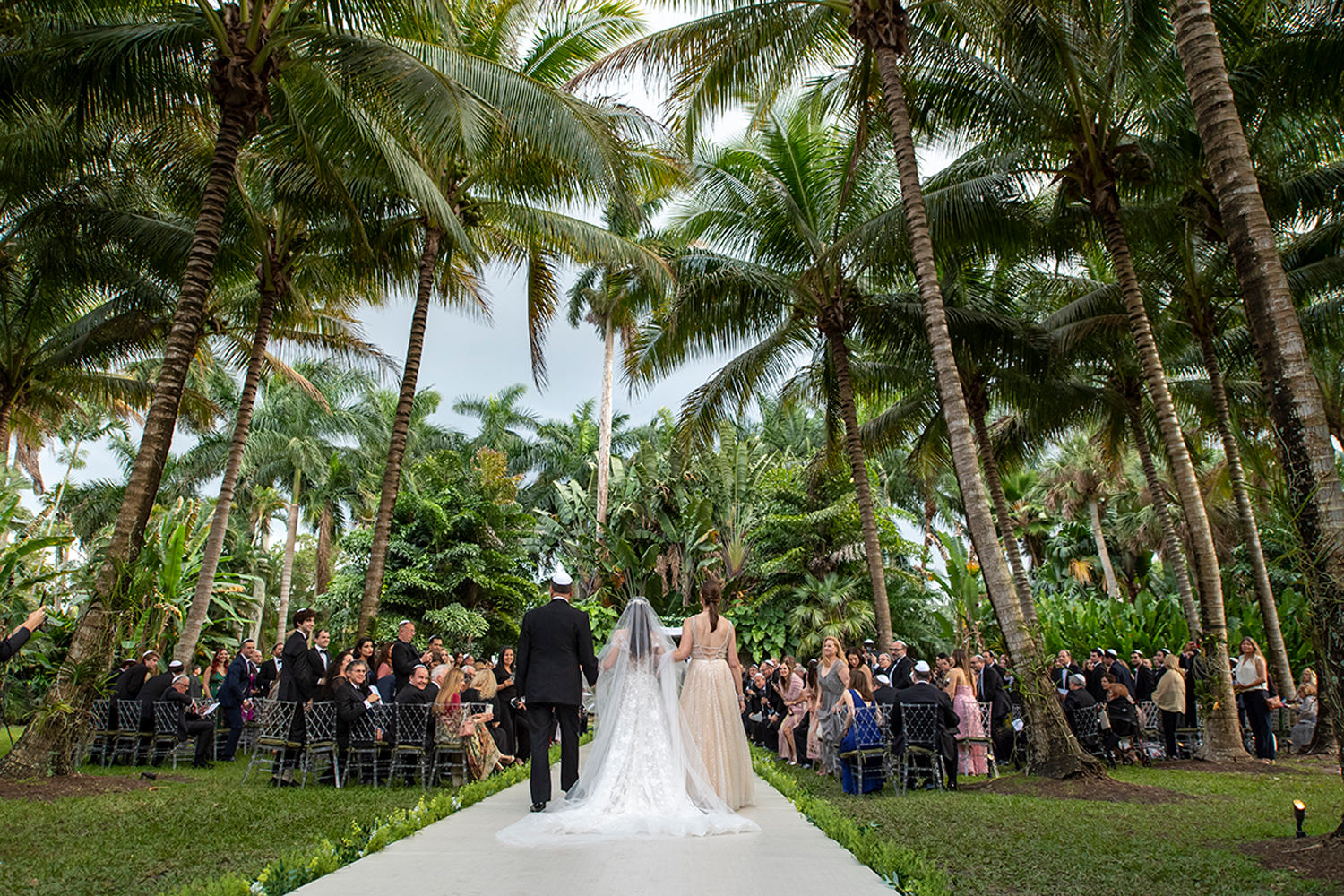 Luxury outdoor wedding ceremony in Miami garden with palm trees, bride walking down aisle with parents, guests seated on both sides, elegant tropical setting