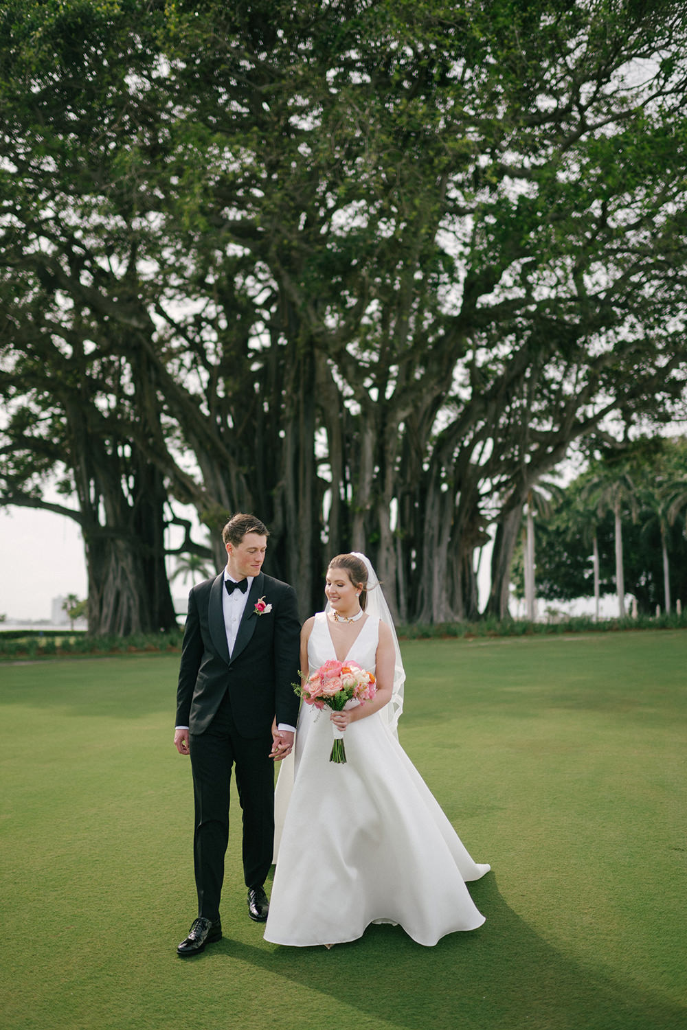 Luxury outdoor wedding ceremony seating in Miami garden with clear chairs, lush greenery, and pastel floral aisle arrangements under banyan trees